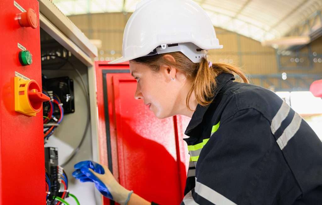 a woman wearing a hard hat and gloves looking at distribution board