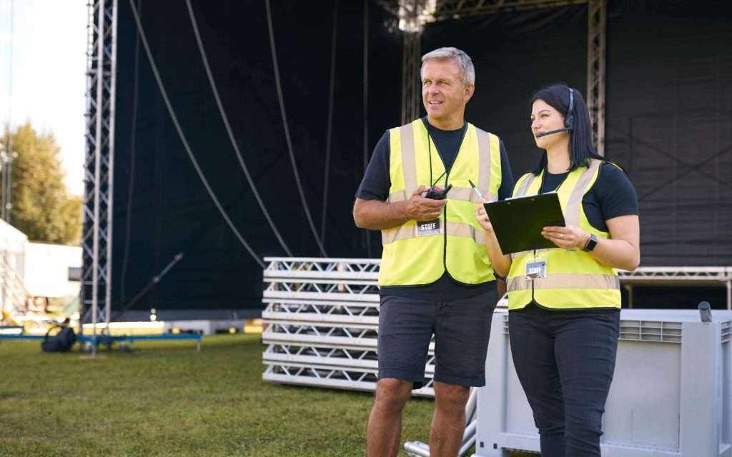Male And Female Production Team With Headsets Setting Up Outdoor Stage For Music Festival Or Concert